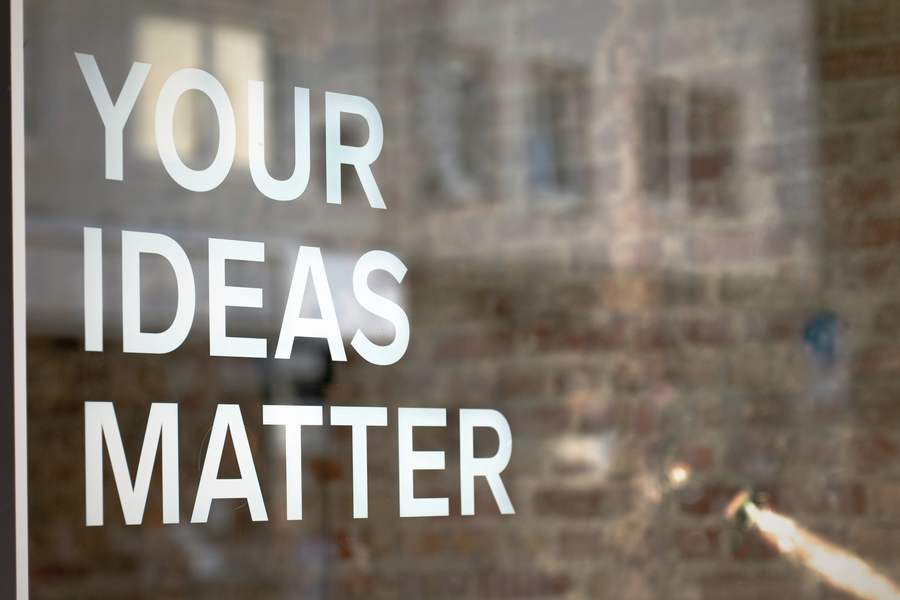 A window sign in white lettering reads Your Ideas Matter, over a warm brick wall, with a soft lens flare at the edge of the frame.